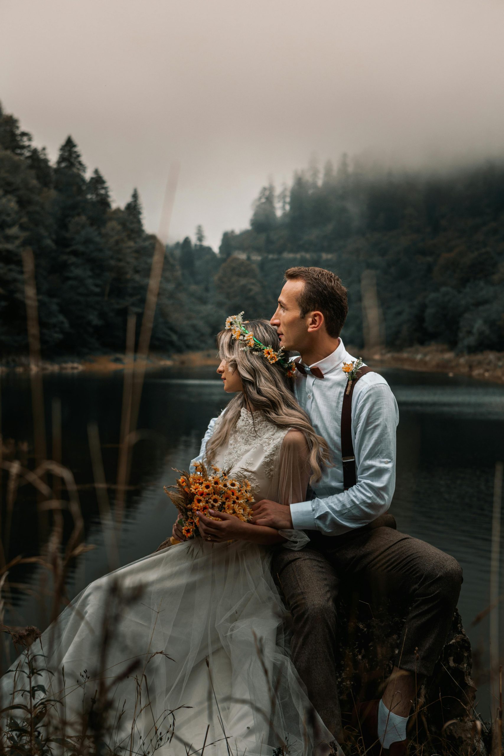 Outdoor destination wedding couple sitting by lake with forest backdrop and bouquet